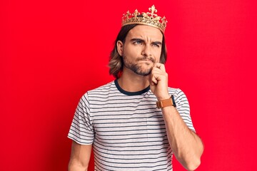 Young handsome man wearing prince crown thinking concentrated about doubt with finger on chin and...