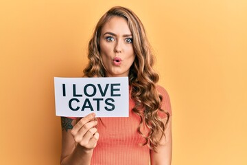 Young blonde girl holding paper with i love cats phrase scared and amazed with open mouth for surprise, disbelief face