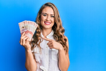 Young blonde girl holding 10 colombian pesos banknotes smiling happy pointing with hand and finger