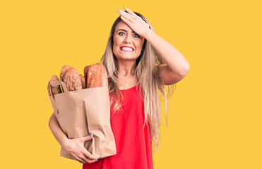 Young beautiful blonde woman holding paper bag with bread stressed and frustrated with hand on head, surprised and angry face