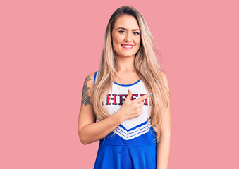 Young beautiful blonde woman wearing cheerleader uniform cheerful with a smile on face pointing...