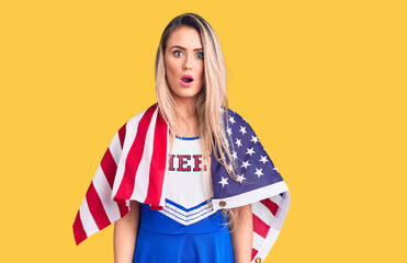 Young beautiful blonde woman wearing cheerleader uniform and united states flag scared and amazed...