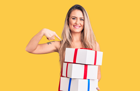 Young beautiful blonde woman holding birthday gifts pointing finger to one self smiling happy and proud