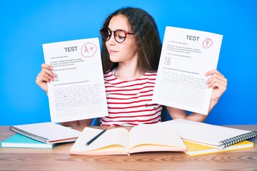 Cute hispanic girl showing failed and passe exam sitting on the table smiling looking to the side and staring away thinking.