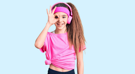 Obraz na płótnie Canvas Cute hispanic child girl wearing gym clothes and using headphones doing ok gesture with hand smiling, eye looking through fingers with happy face.