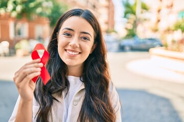 Young hispanic woman smiling happy holding red hiv ribbon walking at the city. - obrazy, fototapety, plakaty