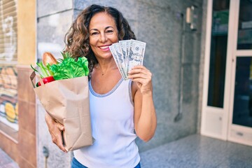 Middle age hispanic woman smiling happy holding a grocery shopping bag full of groceries and american dollars at the city.