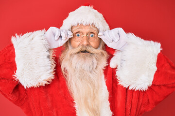 Old senior man with grey hair and long beard wearing santa claus costume holding glasses celebrating crazy and amazed for success with open eyes screaming excited.