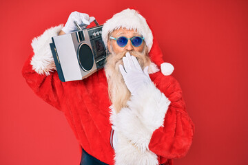 Old senior man wearing santa claus costume and boombox covering mouth with hand, shocked and afraid for mistake. surprised expression