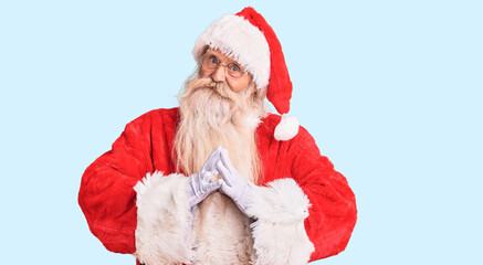 Old senior man with grey hair and long beard wearing traditional santa claus costume hands together...
