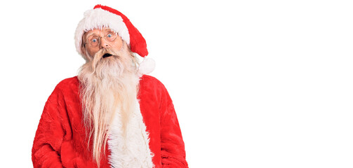 Old senior man with grey hair and long beard wearing traditional santa claus costume in shock face, looking skeptical and sarcastic, surprised with open mouth