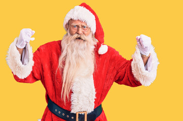 Old senior man with grey hair and long beard wearing traditional santa claus costume angry and mad raising fists frustrated and furious while shouting with anger. rage and aggressive concept.