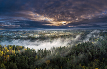 Epic sunrise over the foggy valley in autumn. Morning light lightens colorful forest covered in mist. Impressive storm clouds. 