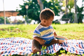 Adorable african american toddler holding hat sitting on the grass at the park.