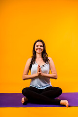 a yoga woman sits with her hands folded Namaste on a purple Mat and looks at the camera on a yellow background