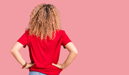 Young blonde woman with curly hair wearing casual red tshirt standing backwards looking away with...