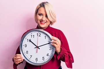 Young beautiful blonde plus size woman doing countdown using big clock over pink background looking positive and happy standing and smiling with a confident smile showing teeth