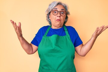 Senior hispanic woman wearing apron and glasses clueless and confused expression with arms and hands raised. doubt concept.