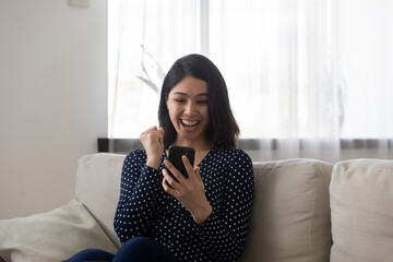 Excited millennial Asian girl relax on sofa in living room look at cellphone screen triumph win...