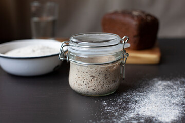 Active rye sourdough starter in a glass jar, a bowl with flour and fresh baked wheat and rye sourdough bread.