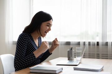 Overjoyed asian young woman sit at desk at home office look at laptop screen triumph win lottery online. Excited happy Vietnamese female feel euphoric read amazing good news or message on computer.