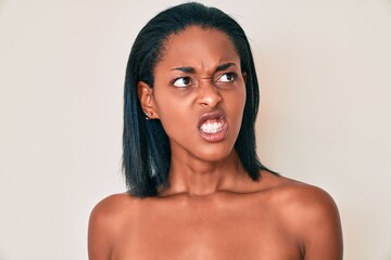 Young african american woman standing topless showing skin angry and mad screaming frustrated and...