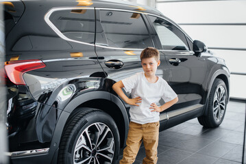 Fototapeta na wymiar At a car dealership, a happy boy stands near a new car before buying it. Car purchase