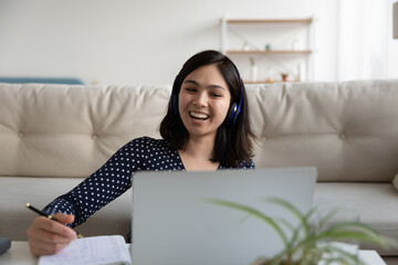 Overjoyed young Asian woman in headphones study distant on laptop laugh watching funny webinar or lesson. Smiling Vietnamese female student have fun write take online course on computer form home.