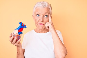 Senior beautiful woman with blue eyes and grey hair holding heart organ serious face thinking about...