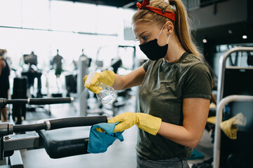 Young female worker disinfecting cleaning and weeping expensive fitness gym equipment with alcohol sprayer and cloth. Coronavirus global world pandemic and health protection safety measures.