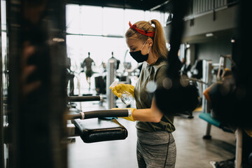 Young female worker disinfecting cleaning and weeping expensive fitness gym equipment with alcohol sprayer and cloth. Coronavirus global world pandemic and health protection safety measures.