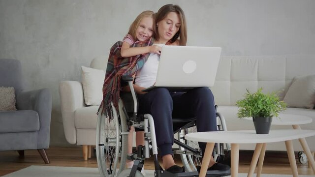 Candid handicapped woman in wheelchair using laptop computer at home, daughter embrace disabled mother, caregiving concept, 