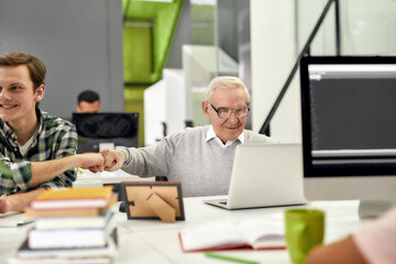 Aged man, senior intern looking at the screen of his laptop and doing a fist bump with his young colleague, Friendly male worker engaging new employee while working in the office
