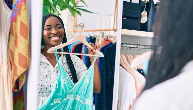 Young african american woman smiling happy holding clothes front of mirror at clothing store