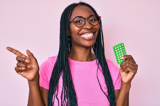 African american woman with braids holding birth control pills smiling happy pointing with hand and finger to the side