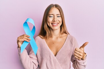 Beautiful young blonde woman holding blue ribbon smiling happy and positive, thumb up doing...