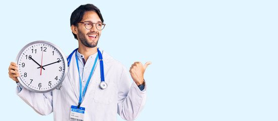 Handsome hispanic man wearing doctor uniform holding clock pointing thumb up to the side smiling...