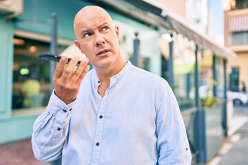 Middle age bald man with serious expression sending audio message using smartphone at the city.