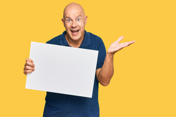 Middle age bald man holding blank empty banner celebrating victory with happy smile and winner expression with raised hands