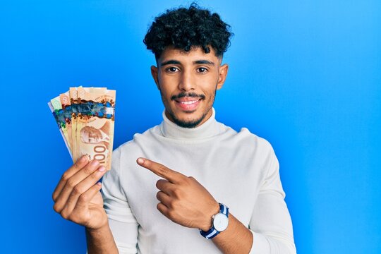 Young arab handsome man holding canadian dollars smiling happy pointing with hand and finger