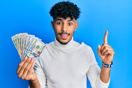 Young arab handsome man holding dollars smiling with an idea or question pointing finger with happy face, number one