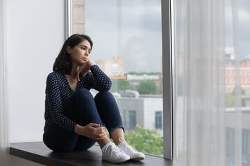 Sad young asian woman sit on windowsill at home look in window distance think feel lonely lack...