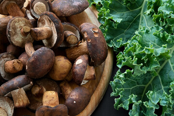 Brown wild mushrooms on wooden cutting board and green kale leaves on table, top view, backgrounds

