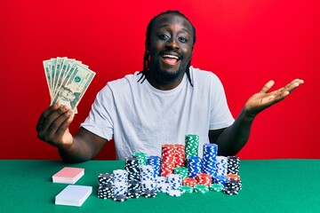 Handsome young black man playing poker holding 20 dollars banknotes celebrating achievement with happy smile and winner expression with raised hand
