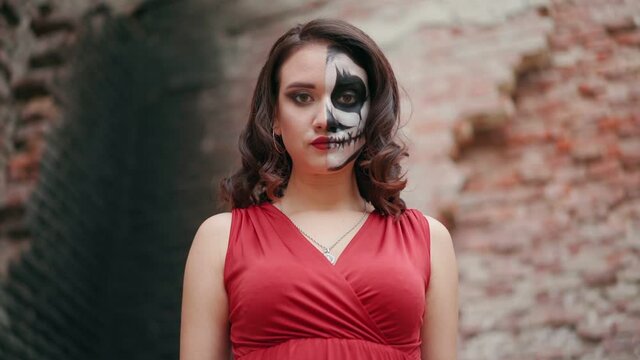 Portrait beautiful woman in a red evening dress and awesome Halloween makeup stands against the backdrop of an abandoned building. Greasepaint for the Holiday on October 31. Slow motion