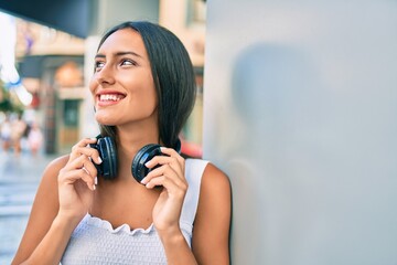 Young latin girl smiling happy using headphones leaning on the wall.