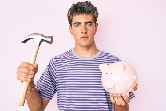 Young Handsome Man Holding Piggy Bank And Hammer Relaxed With Serious Expression On Face. Simple And Natural Looking At The Camera.