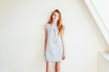 Happy teenage girl smiling. Portrait young happy positive teen woman in grey dress at home in bright room against white wall. European woman. Positive human emotion facial expression body language.