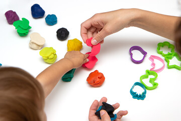 Toddler sculpts from colored plasticine on a white table. The hand of a small child squeezes pieces of colored plasticine.