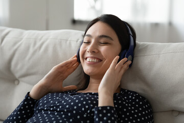 Happy millennial asian girl in wireless modern headphones relax on sofa at home listen to music. Smiling young Vietnamese woman in earphones rest on couch enjoy good quality sound on gadget.